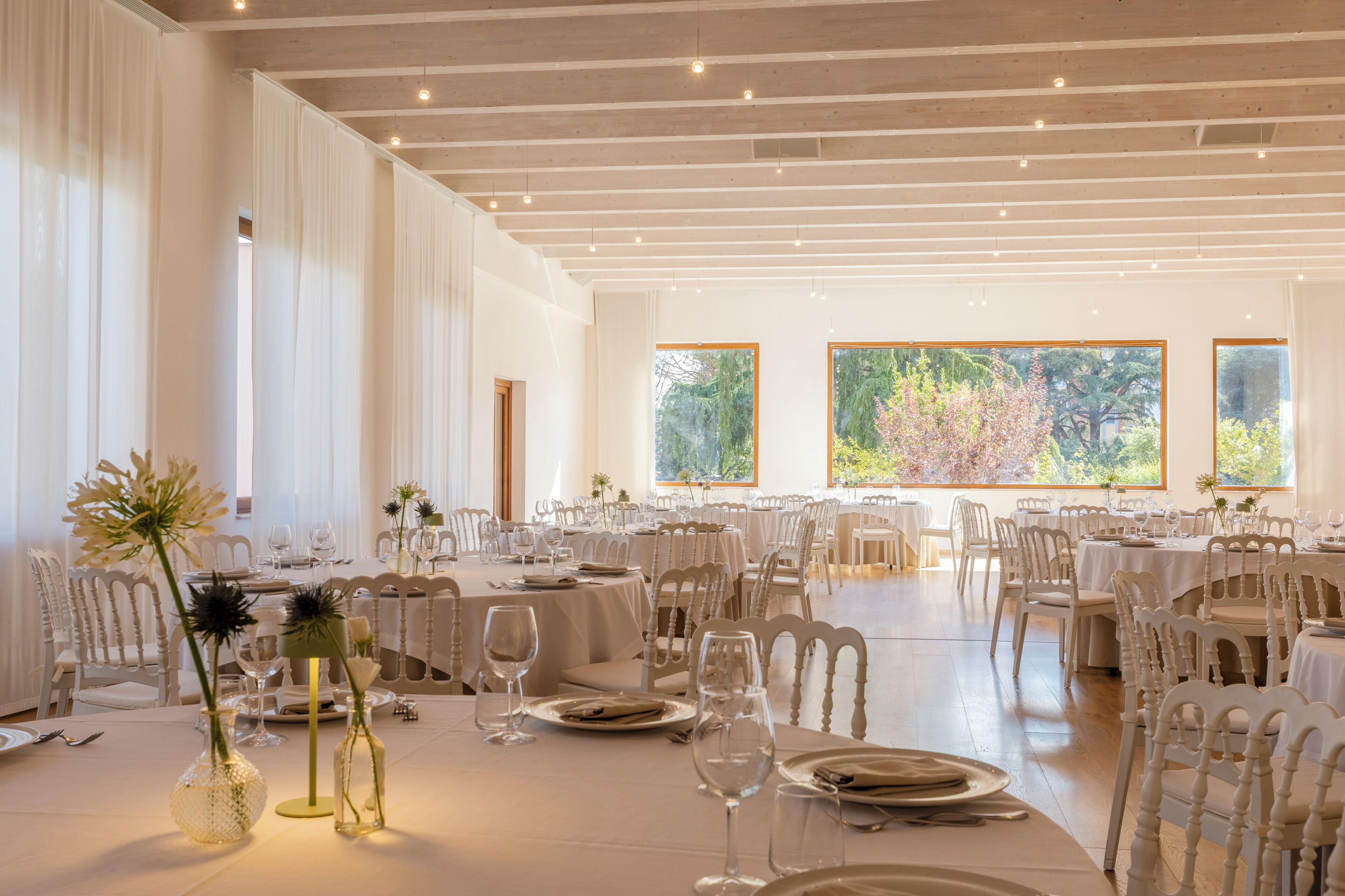 A spacious dining hall with multiple tables set for a meal, large windows offering a view of greenery, and a high ceiling with exposed beams.