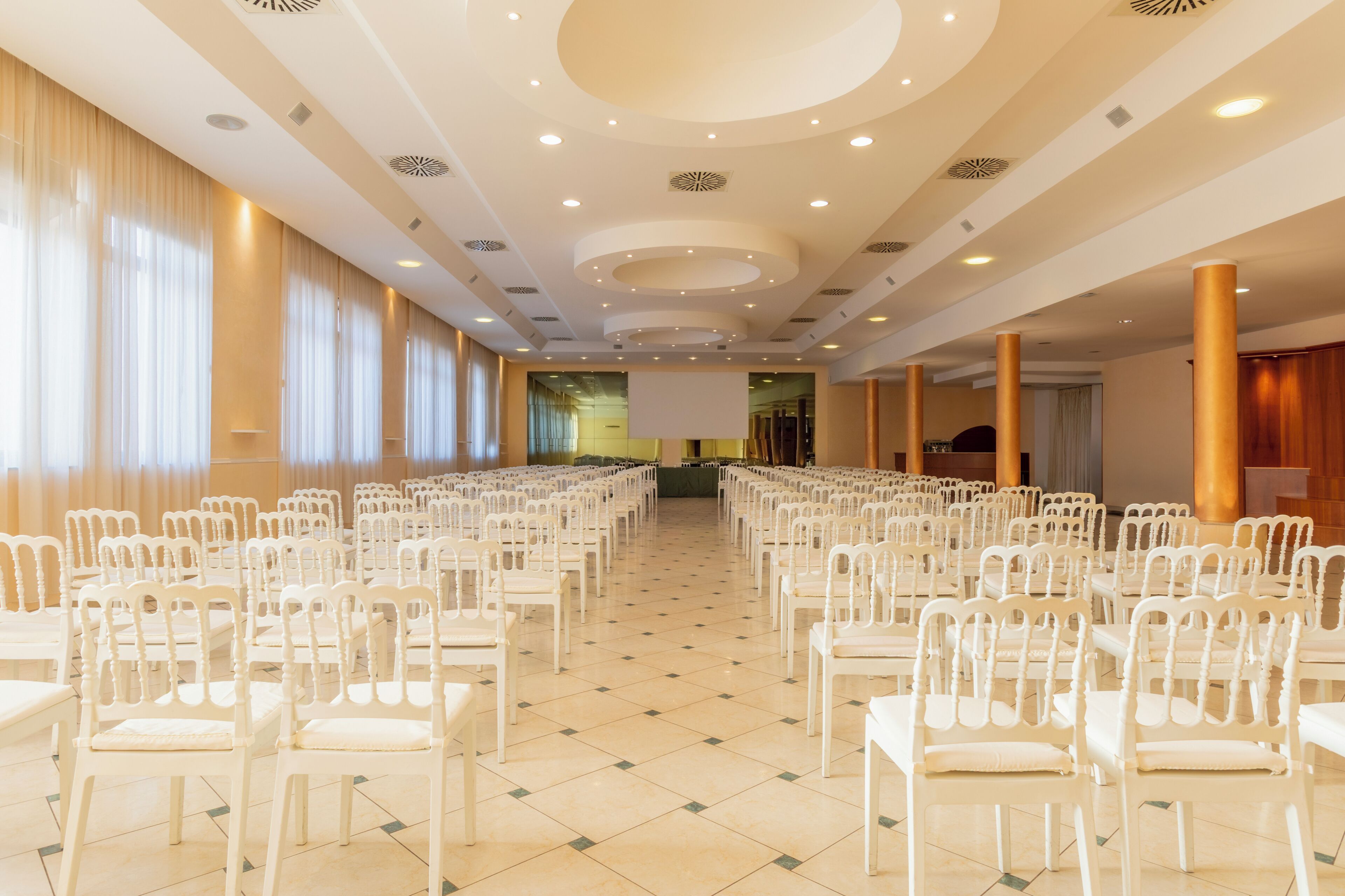 Un salón de conferencias con filas de sillas blancas, piso de baldosas y un diseño moderno en el techo.