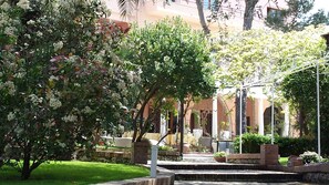 Un patio con varios árboles y arbustos, escalones de piedra y un edificio con arcos al fondo.