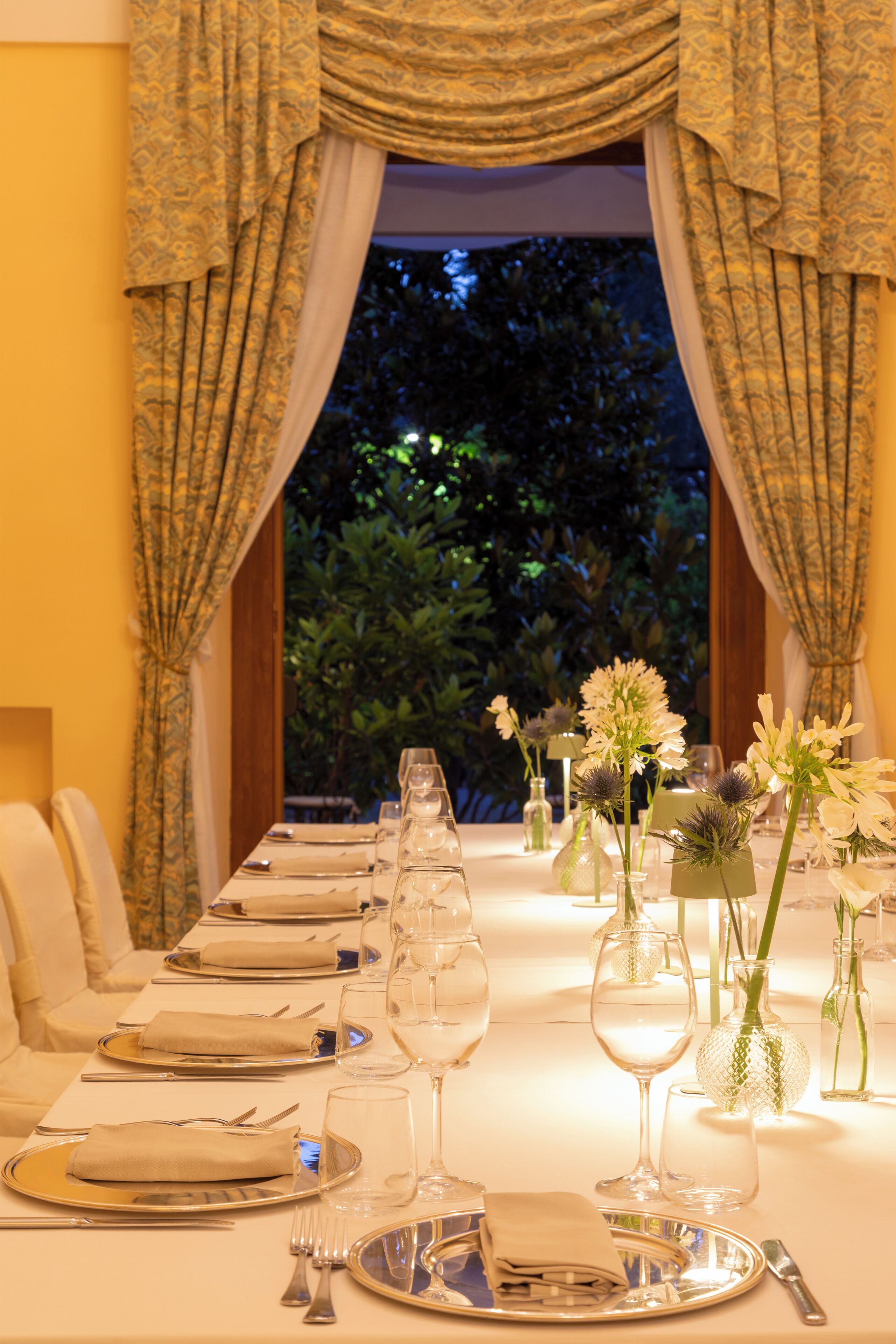 Una mesa de comedor formal dispuesta con varias copas de vino, platos y cubiertos, con vista a zonas verdes al exterior.