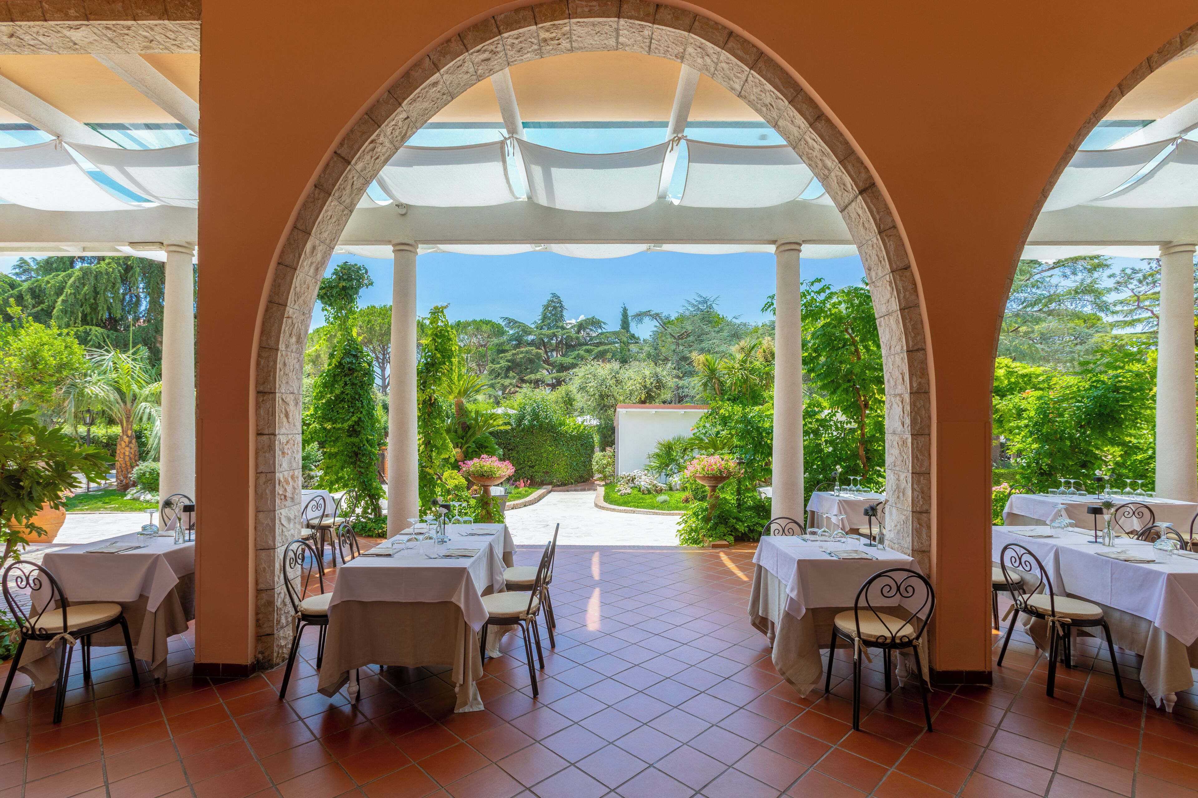 Un área de comedor al aire libre techada, con mesas y sillas, vistas a un jardín y cielo despejado.