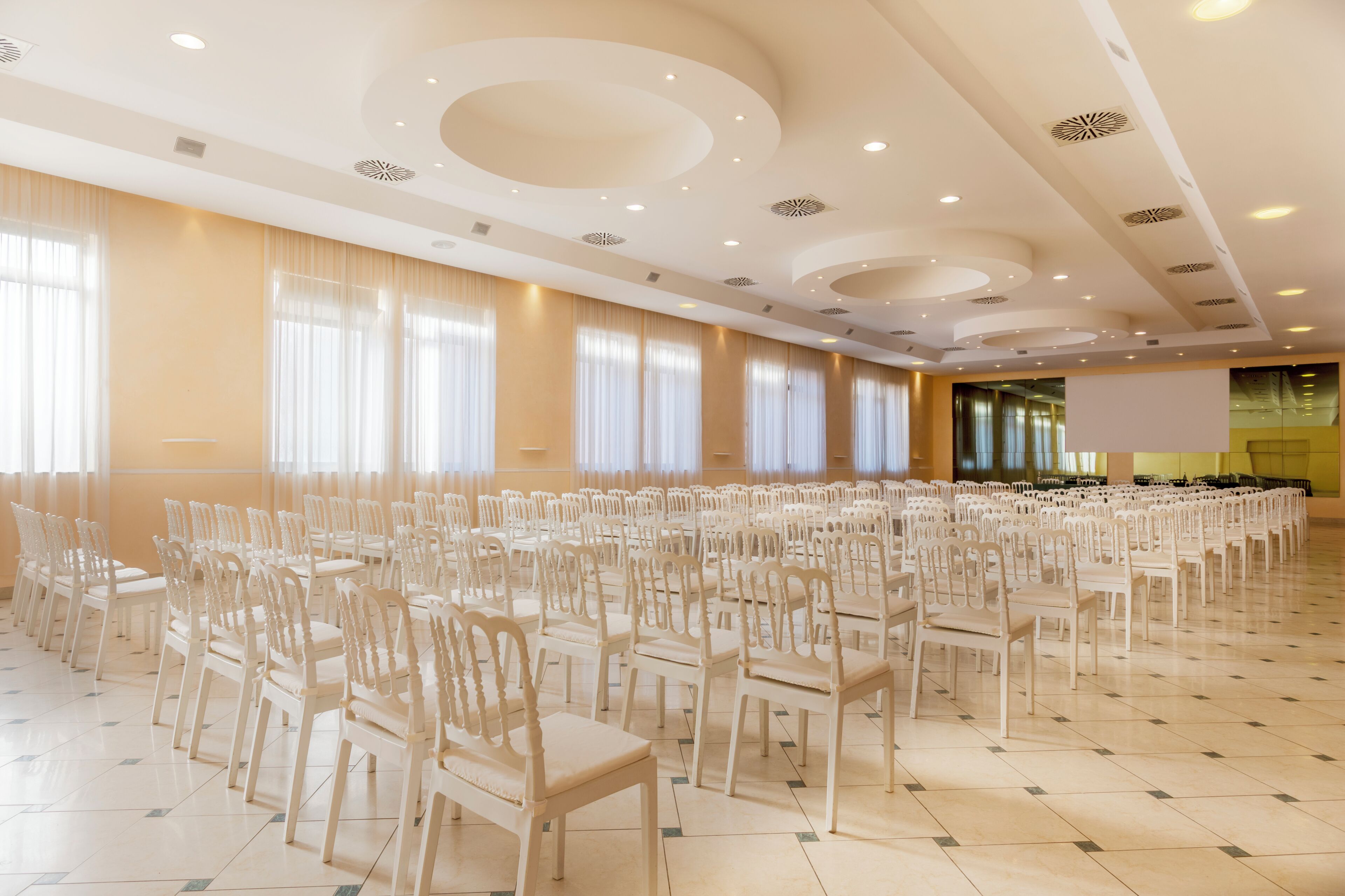 Un salón amplio con filas de sillas blancas, un techo alto con lámparas circulares y ventanas grandes con cortinas traslúcidas.