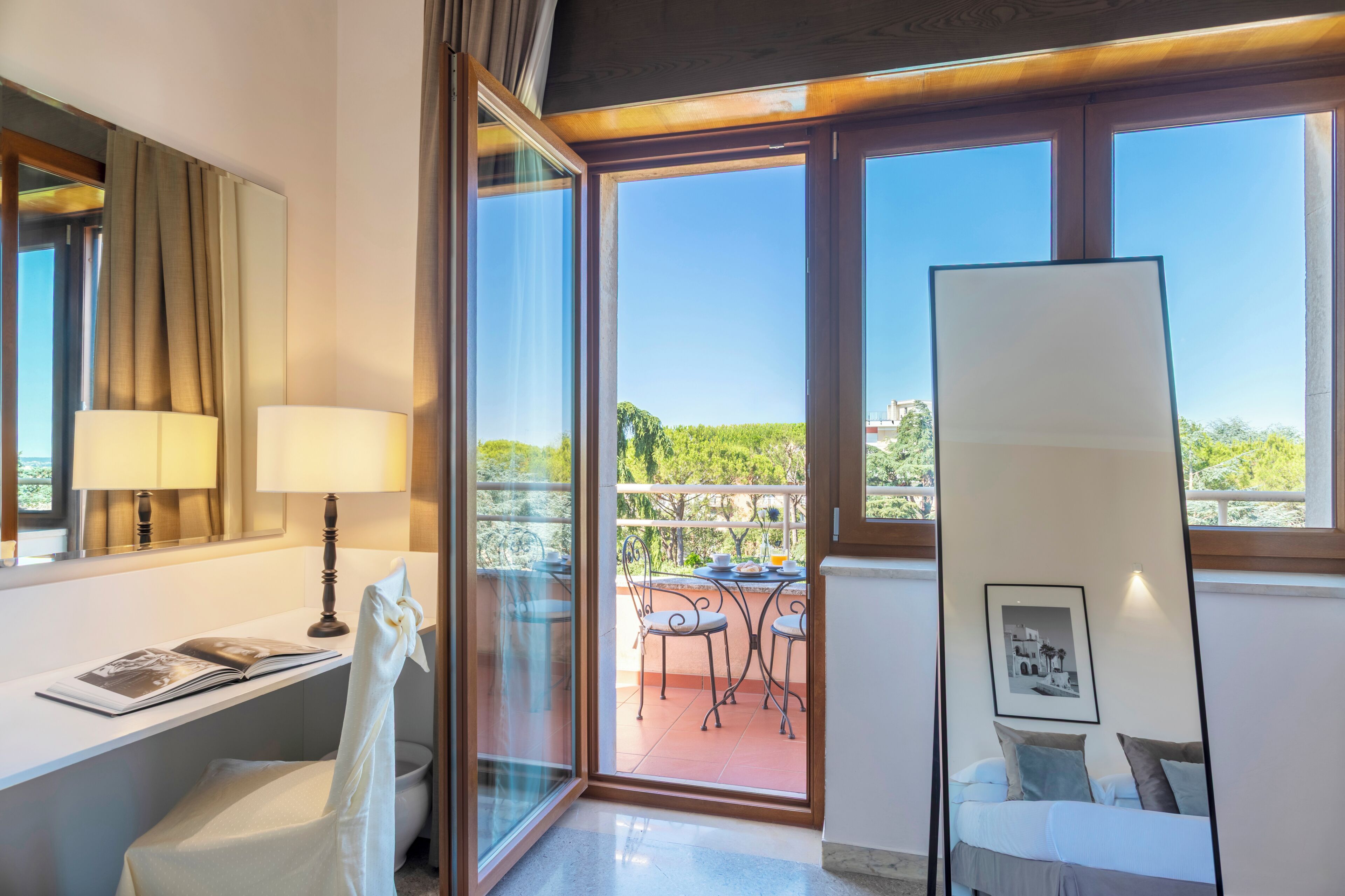 A hotel room with a balcony, a desk with a lamp, a bed with pillows, and a framed picture on the wall.
