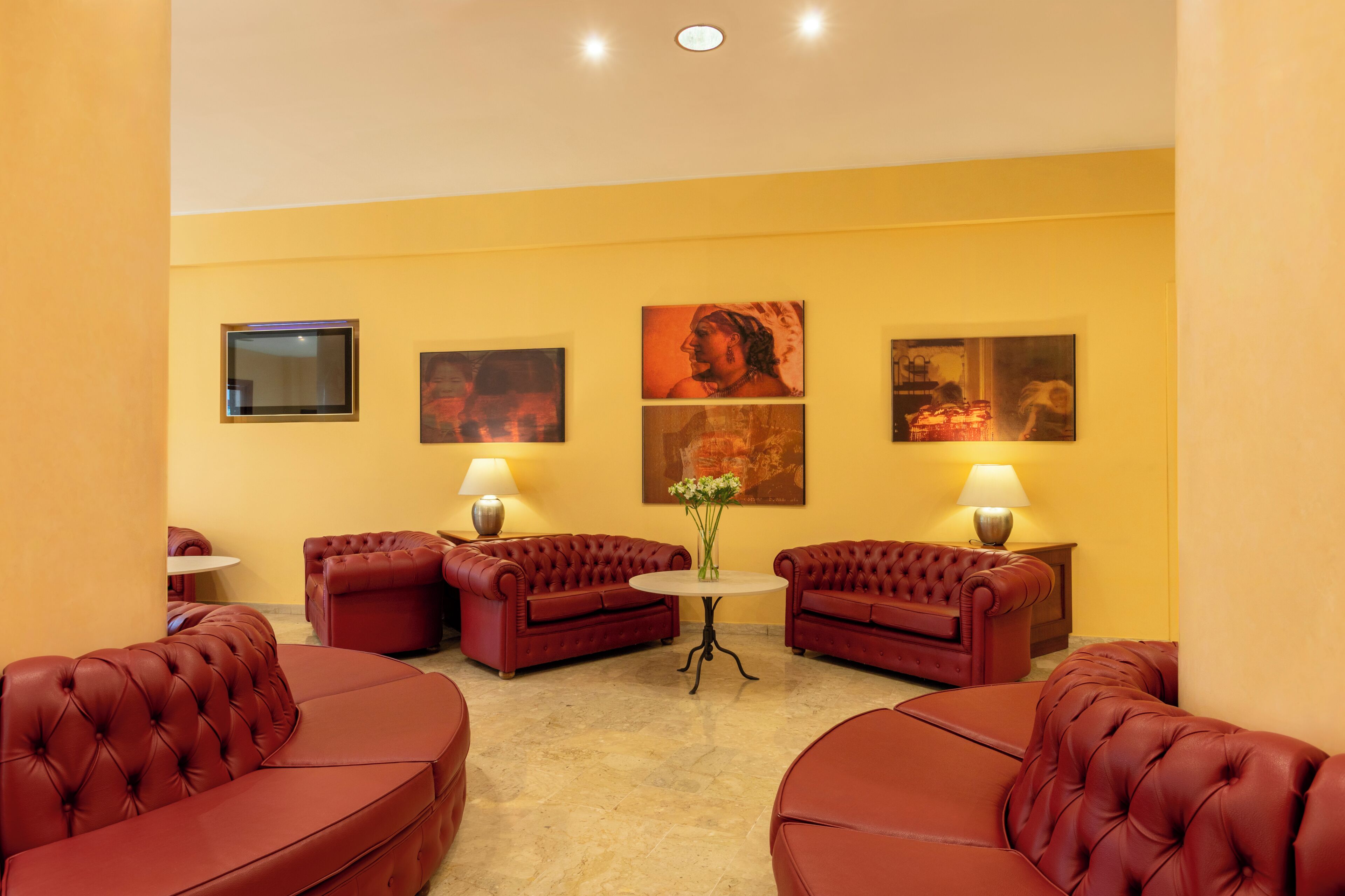Habitación de hotel con pared amarilla, dos sofás de cuero rojo, una mesa redonda pequeña y tres cuadros colgados en la pared.