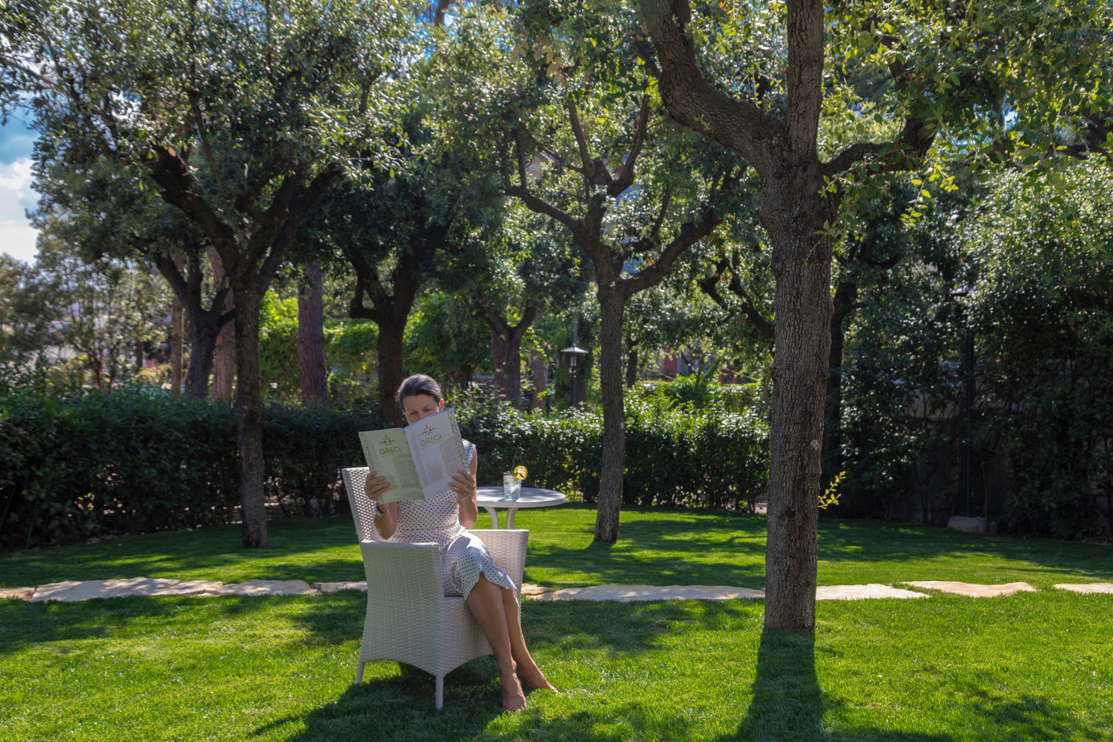 A person seated on a wicker chair in a grassy outdoor area with trees and a table with a glass and a book.