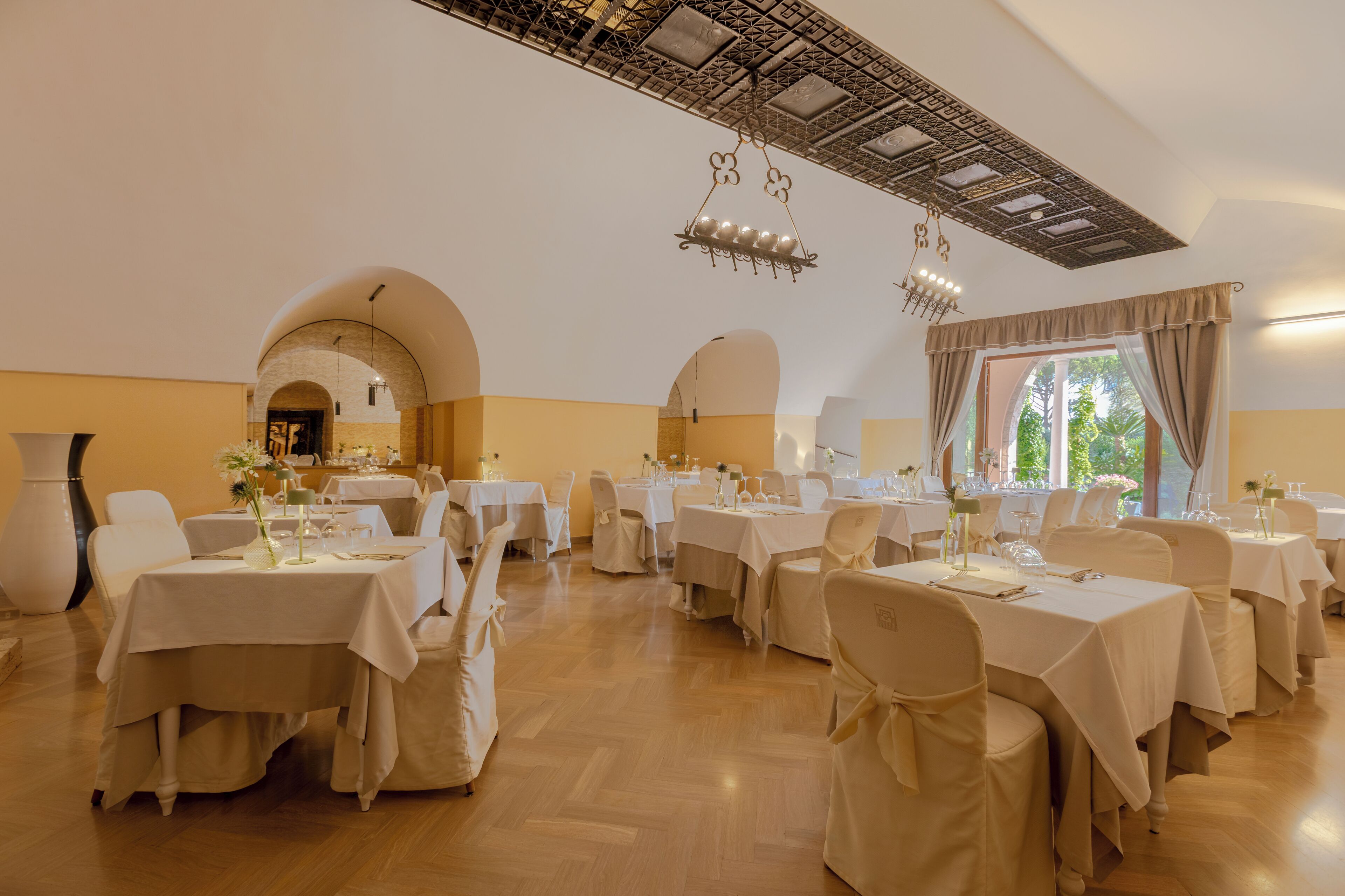A spacious dining hall with multiple tables set for a meal, featuring a high ceiling with intricate metalwork and large windows allowing natural light.