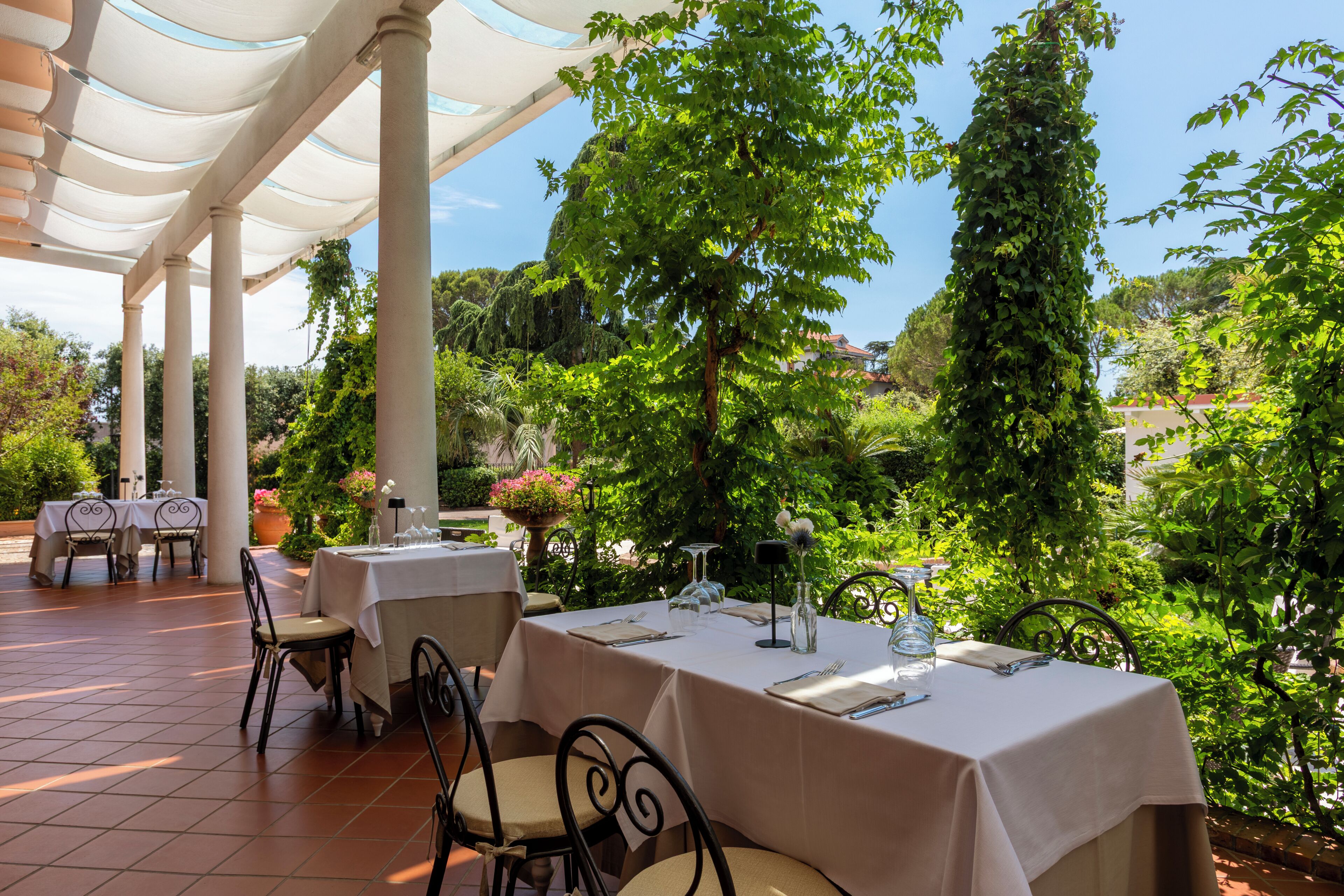 An outdoor dining area with tables set for two, surrounded by greenery and a clear sky.