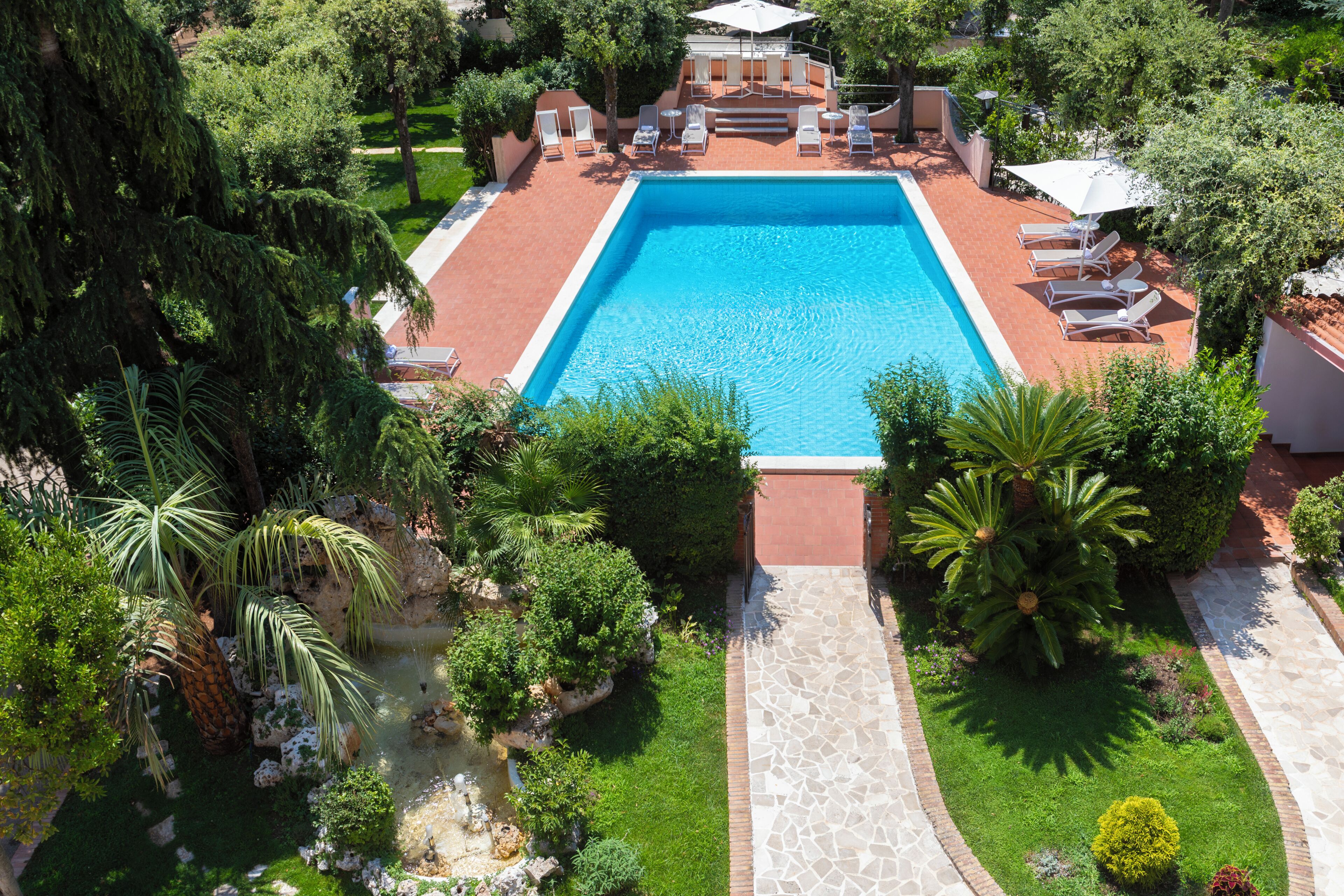 A rectangular swimming pool with a tiled deck, surrounded by lounge chairs and umbrellas, with a garden and trees in the background.