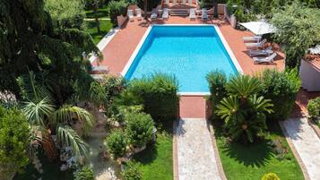 A rectangular swimming pool with a tiled deck, surrounded by lounge chairs and umbrellas, with a garden and trees in the background.