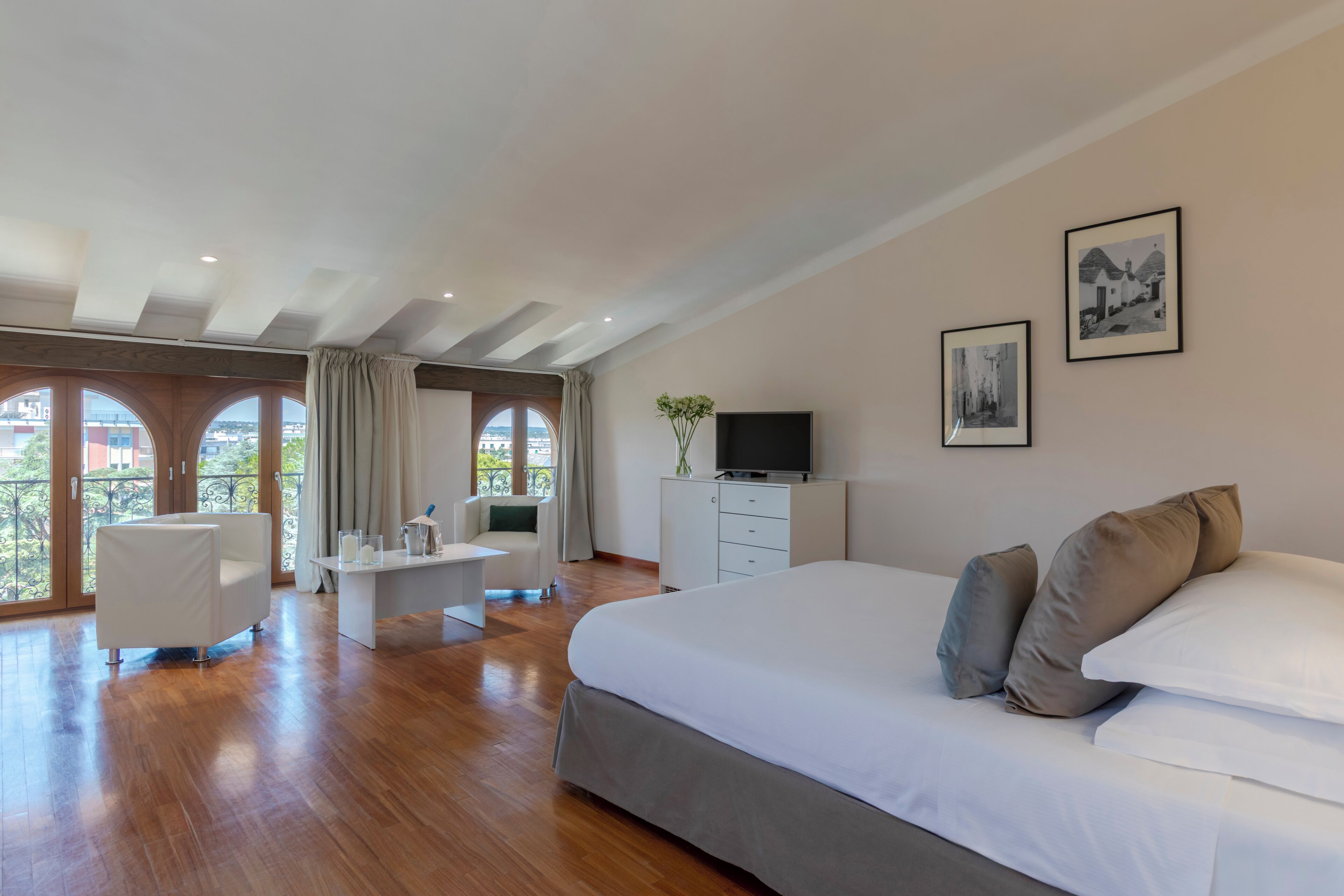 Une chambre spacieuse avec un grand lit, un bureau et une chaise. Elle comprend deux fenêtres avec des rideaux, un téléviseur posé sur une commode et des œuvres d’art accrochées au mur.