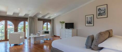 Une chambre spacieuse avec un grand lit, un bureau et une chaise. Elle comprend deux fenêtres avec des rideaux, un téléviseur posé sur une commode et des œuvres d’art accrochées au mur.