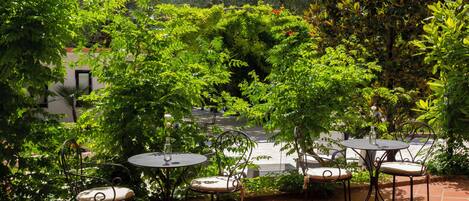 An outdoor patio area with round tables and chairs, surrounded by lush greenery and trees.