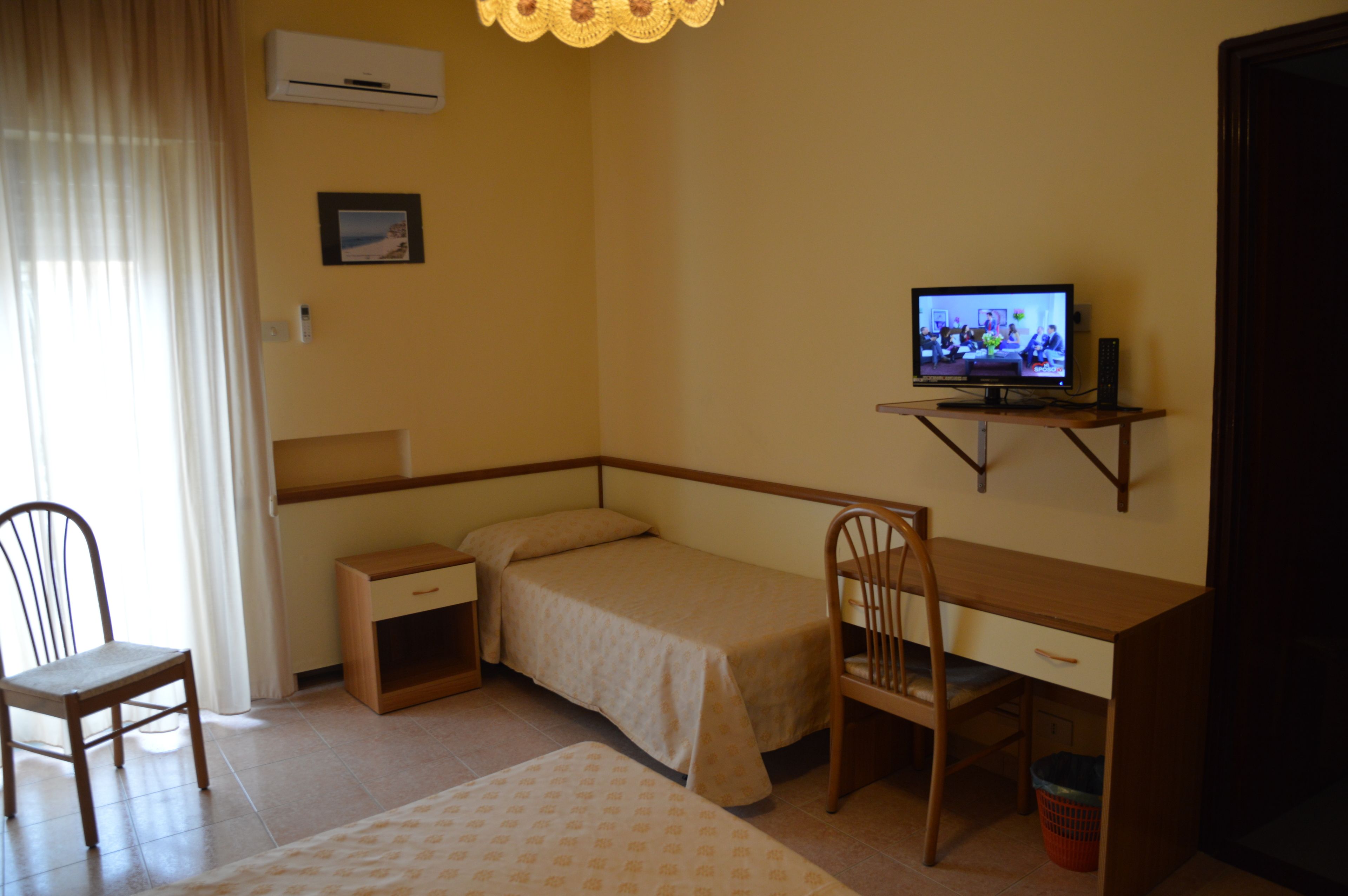 Une petite chambre à coucher, bien rangée, avec un lit simple, un bureau, une chaise, un téléviseur et une petite table.