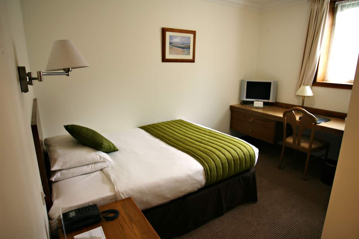 Quarto de hotel com cama, escrivaninha, cadeira, televisão e luminária.