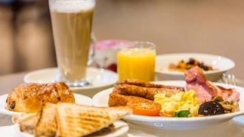 Une table dressée pour le petit-déjeuner avec des croissants, des œufs brouillés, des saucisses, du jambon et un verre de jus d’orange.