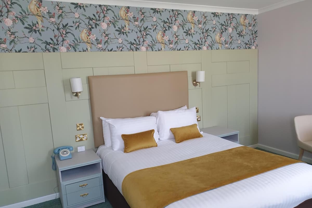 Cama bem arrumada com almofadas brancas e mostarda, cabeceira marrom e papel de parede com padrão floral.