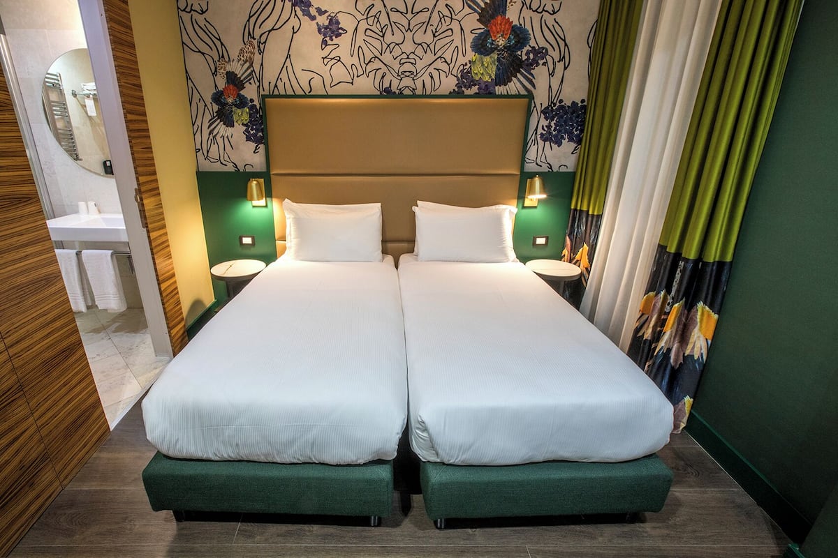 A double bed with two pillows, a nightstand with a lamp, a mirror, and a bathroom with a sink and towels.