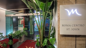 A modern lobby with a glass elevator, potted plants, and a sign for Roma Centro St. John. - Mercure Roma Centro St. John (Rome)