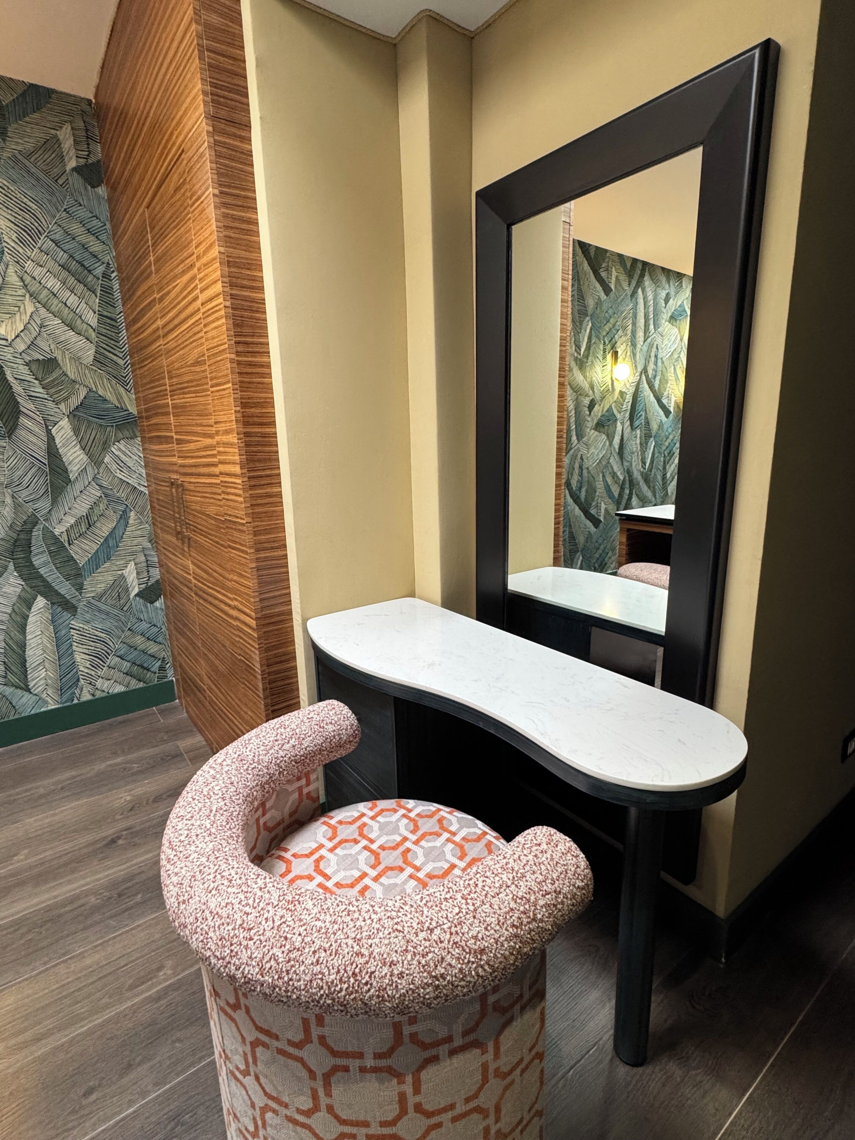 A bathroom with a vanity, a mirror, a patterned stool, and a decorative wall.
