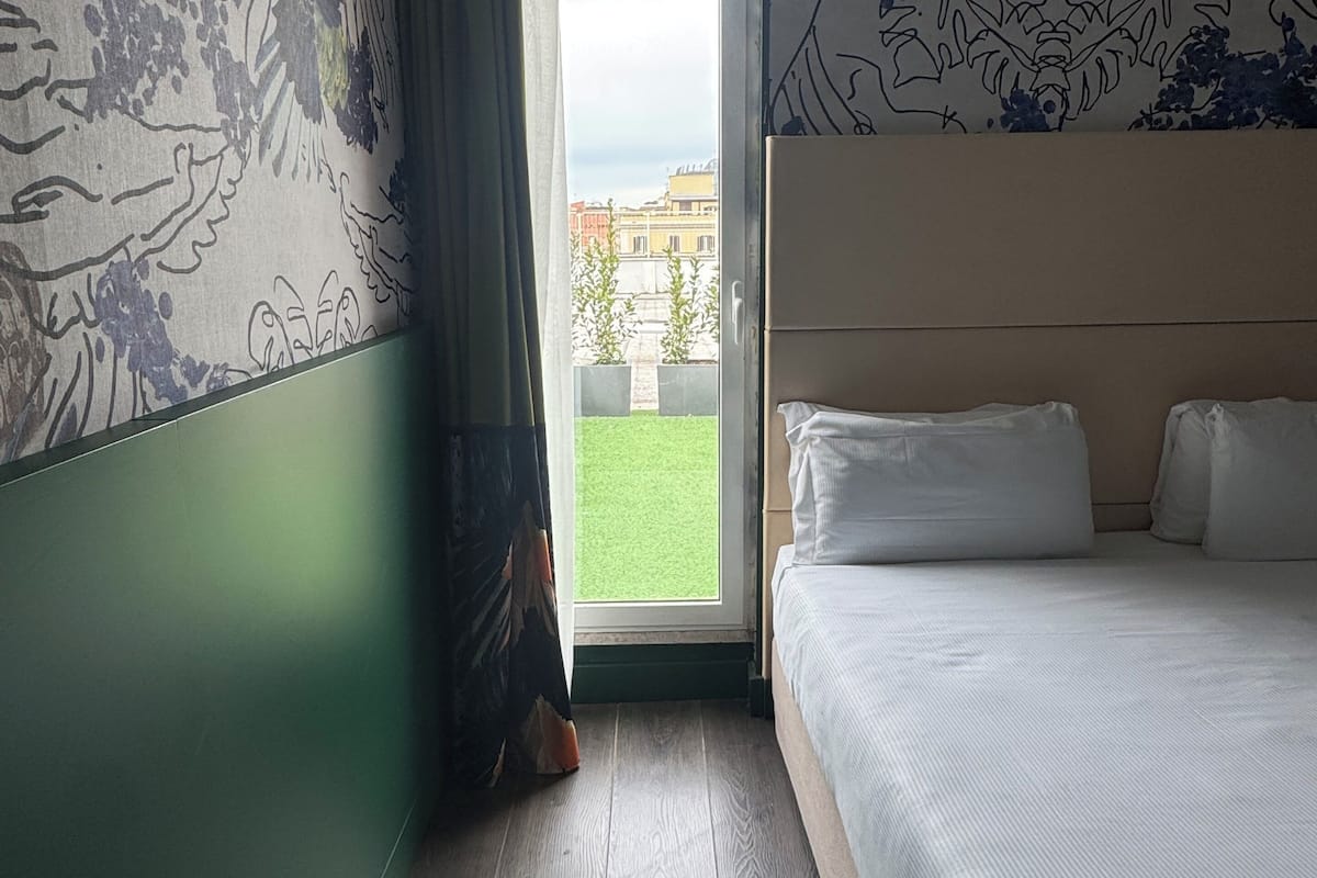 A hotel room with a bed, a window with a view of buildings, and a door.