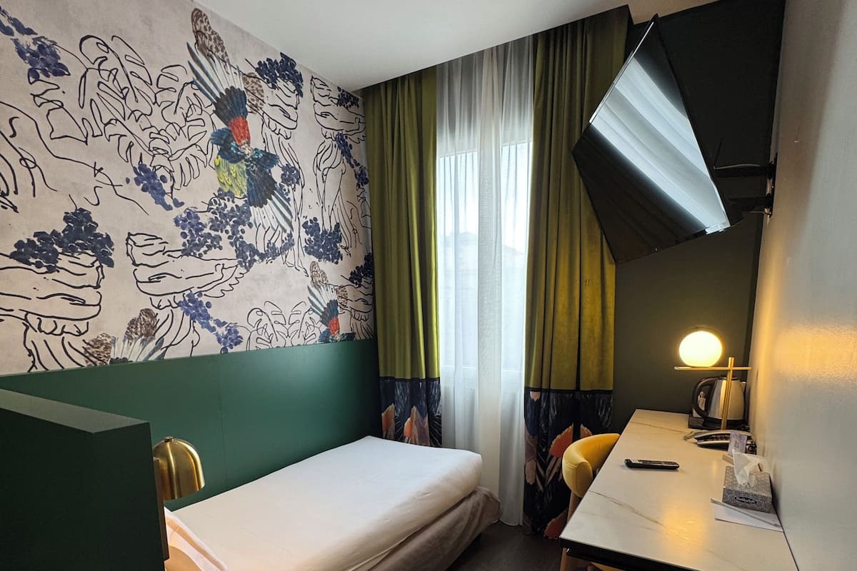 A compact hotel room with a single bed, a desk with a lamp, a chair, and a television mounted on the wall.