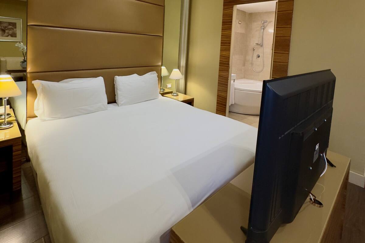A hotel room with a bed, bedside lamps, a TV, a wooden headboard, a mirror, and a shower area.