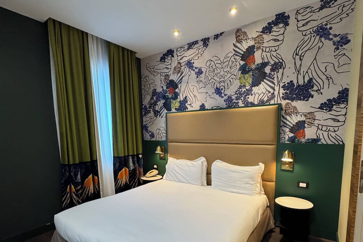 A hotel room with a large bed, a bedside table, a nightstand, a wall-mounted lamp, and a patterned wall.