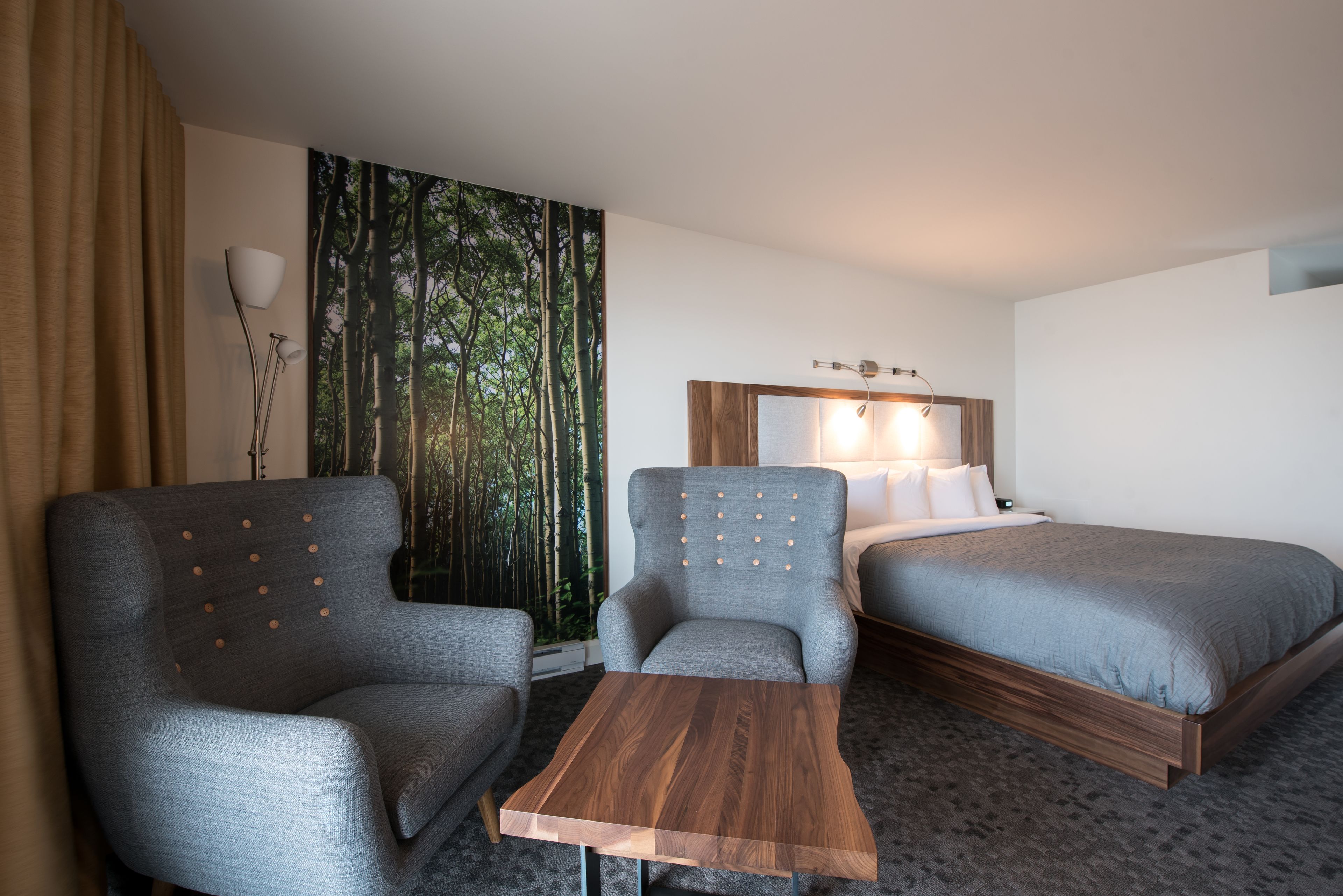 A hotel room with two grey armchairs, a wooden coffee table, a bed with a wooden headboard, and a large window with a forest view.