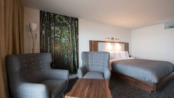 A hotel room with two grey armchairs, a wooden coffee table, a bed with a wooden headboard, and a large window with a forest view.