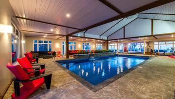 An indoor swimming pool with red lounge chairs, a stone floor, and large windows.