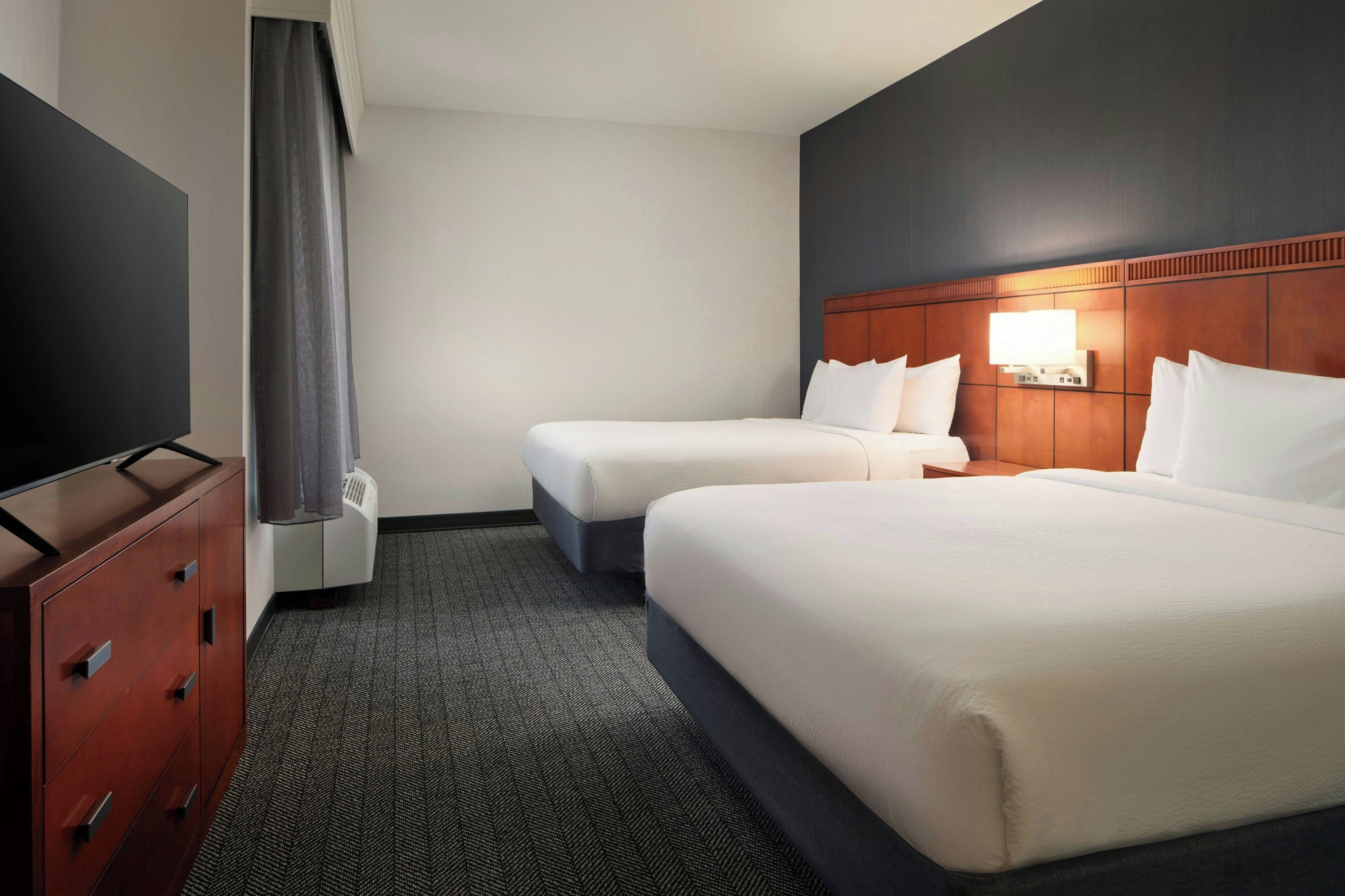 A hotel room with two beds, a TV, a dresser, and a wall-mounted light.