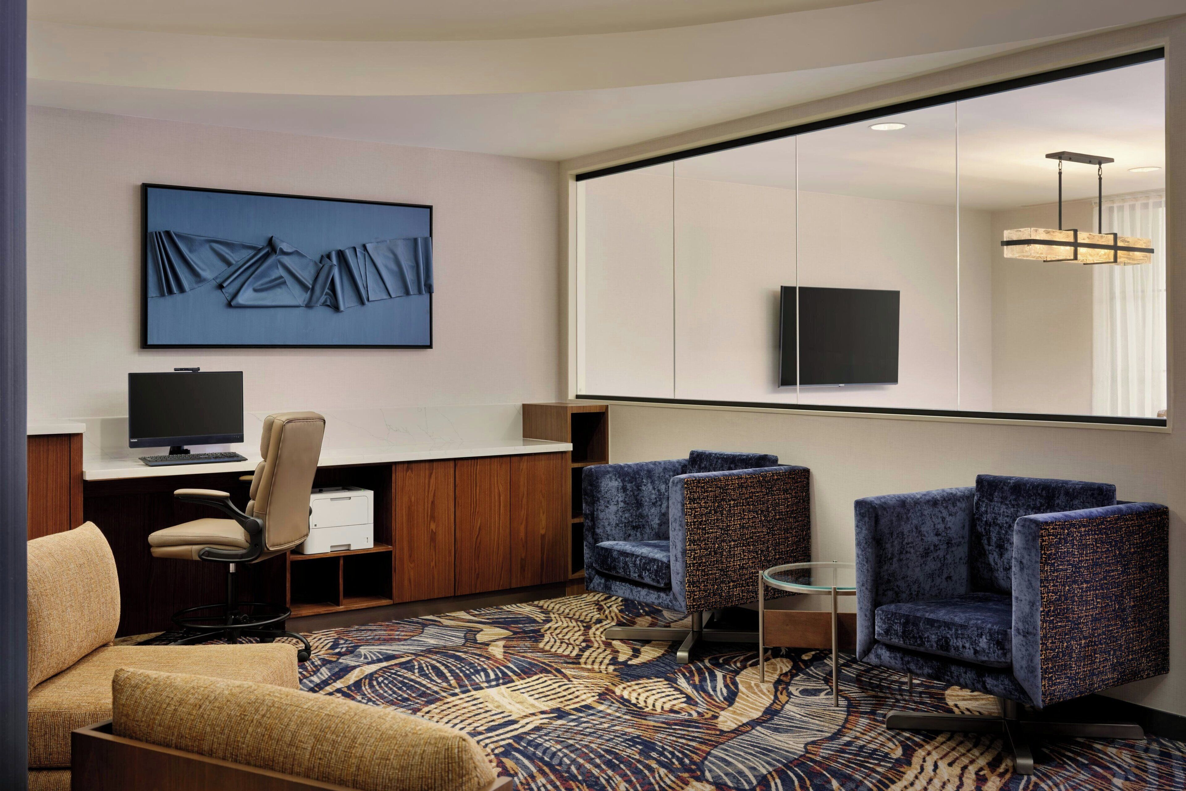 Een hotelkamer met een grote spiegel, een bureau met een computer, twee stoelen, een bank en een kamerbreed tapijt met een patroon.