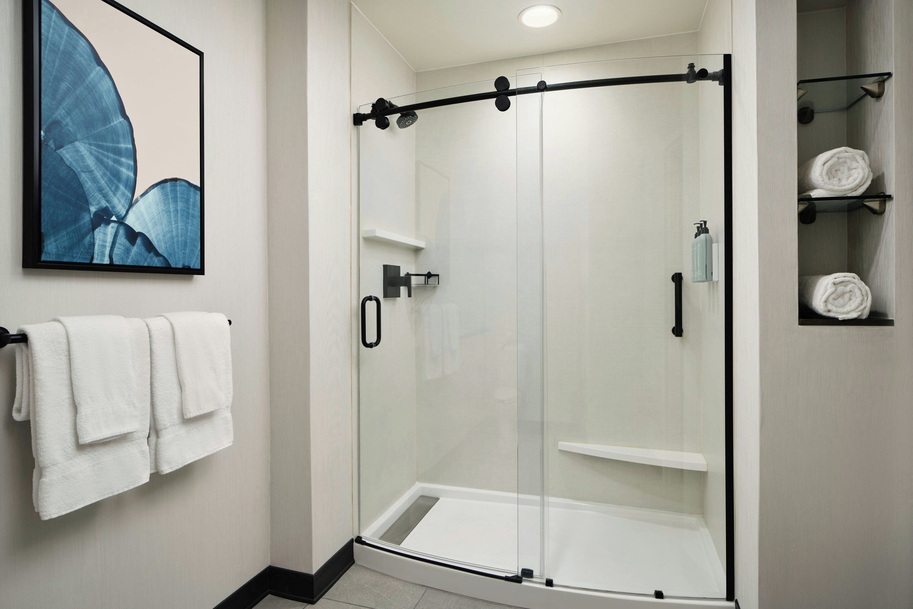 A glass-enclosed shower with a black handle, a white towel rack with towels, and a framed artwork on the wall.