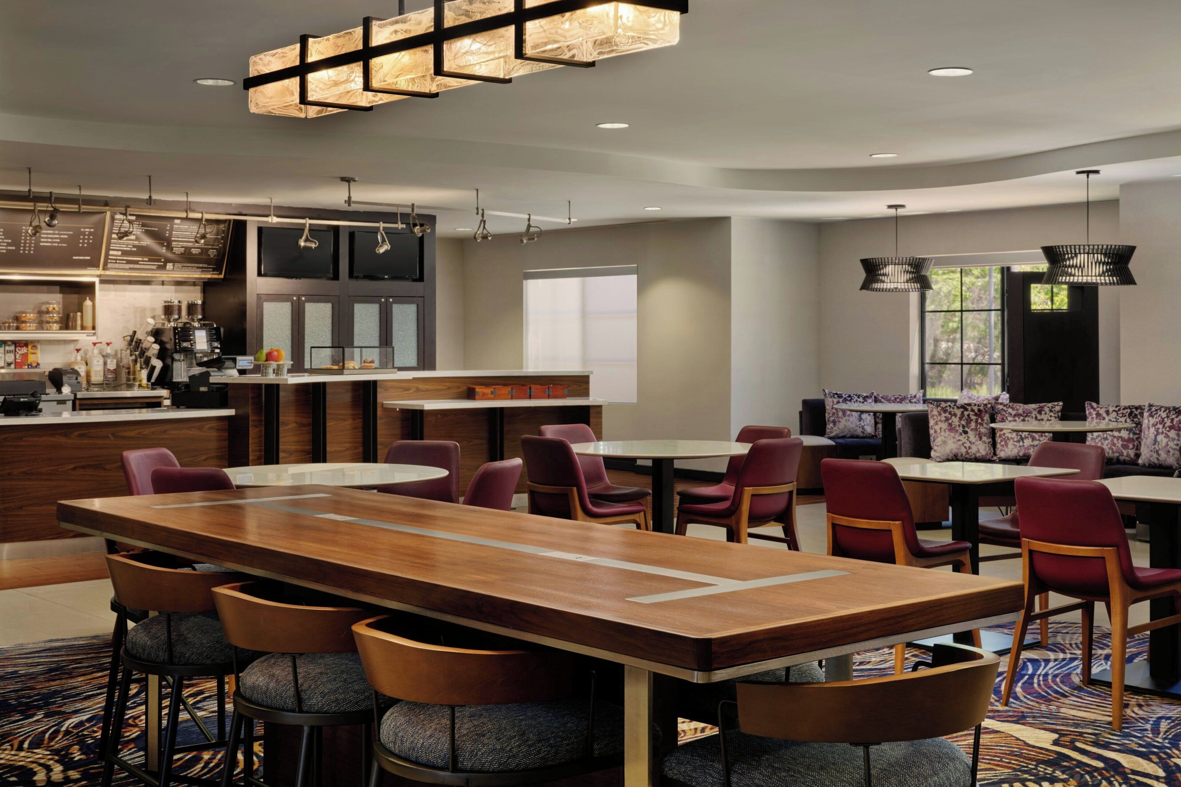 Una moderna hall d'albergo con una lunga reception in legno, diverse zone relax e una cucina a vista.
