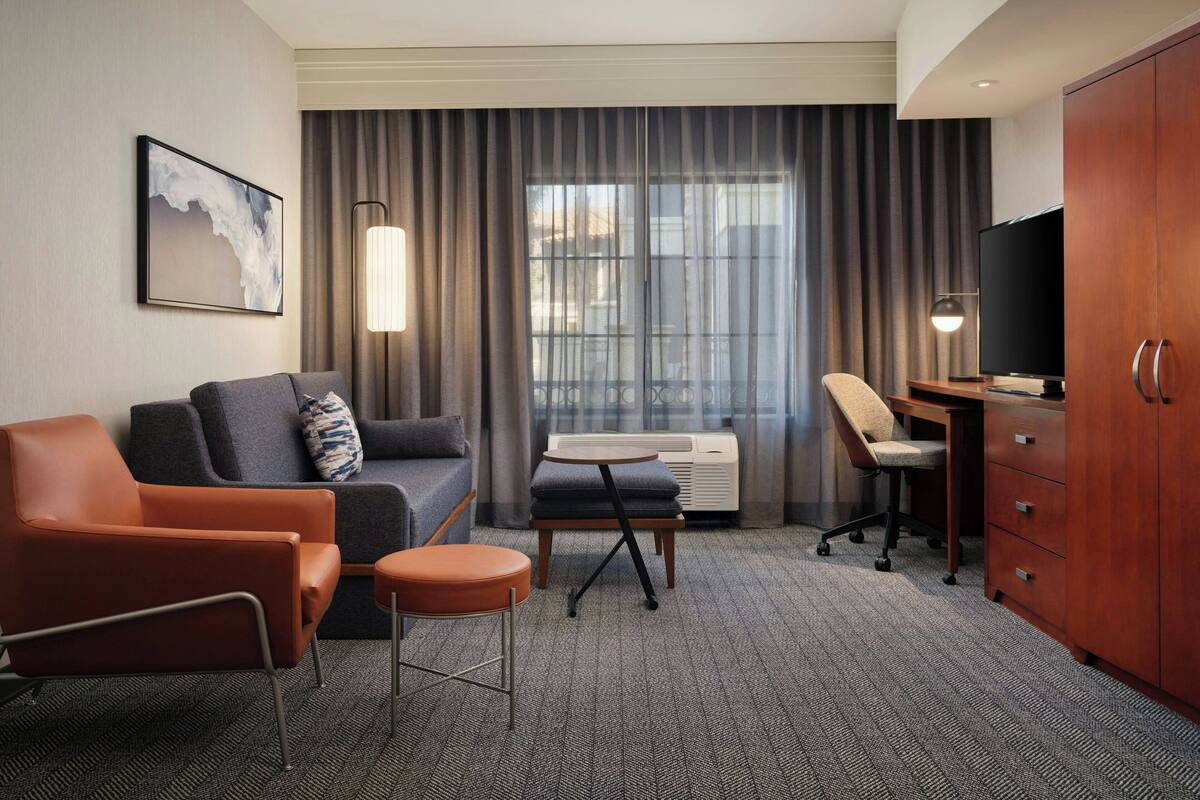 Une chambre d’hôtel équipée d’un canapé, d’un fauteuil, d’un bureau, d’une télévision et d’une grande fenêtre avec des rideaux.