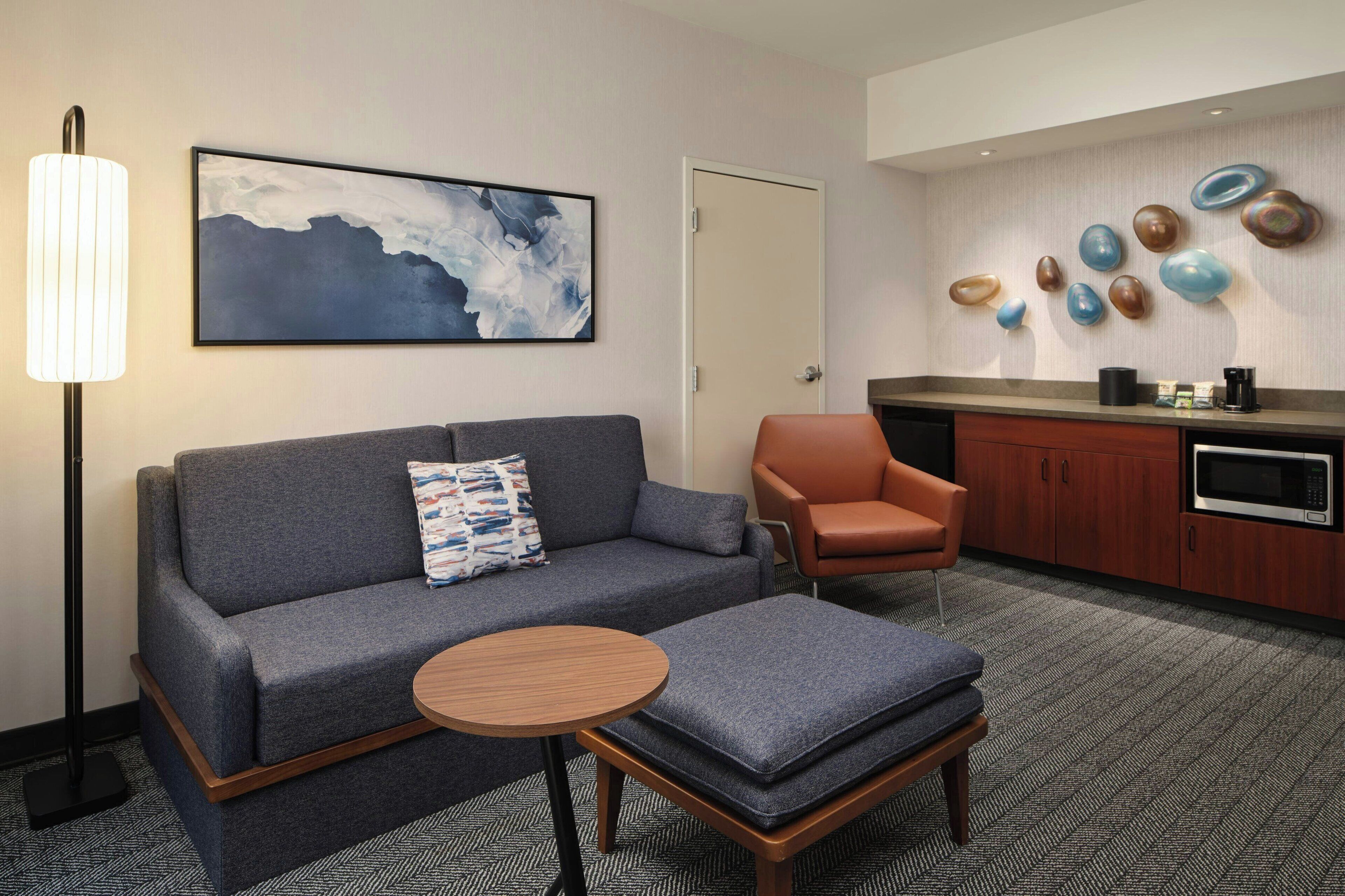 A modern hotel room with a gray sofa, a wooden side table, a small orange chair, a microwave, and a kitchen counter.