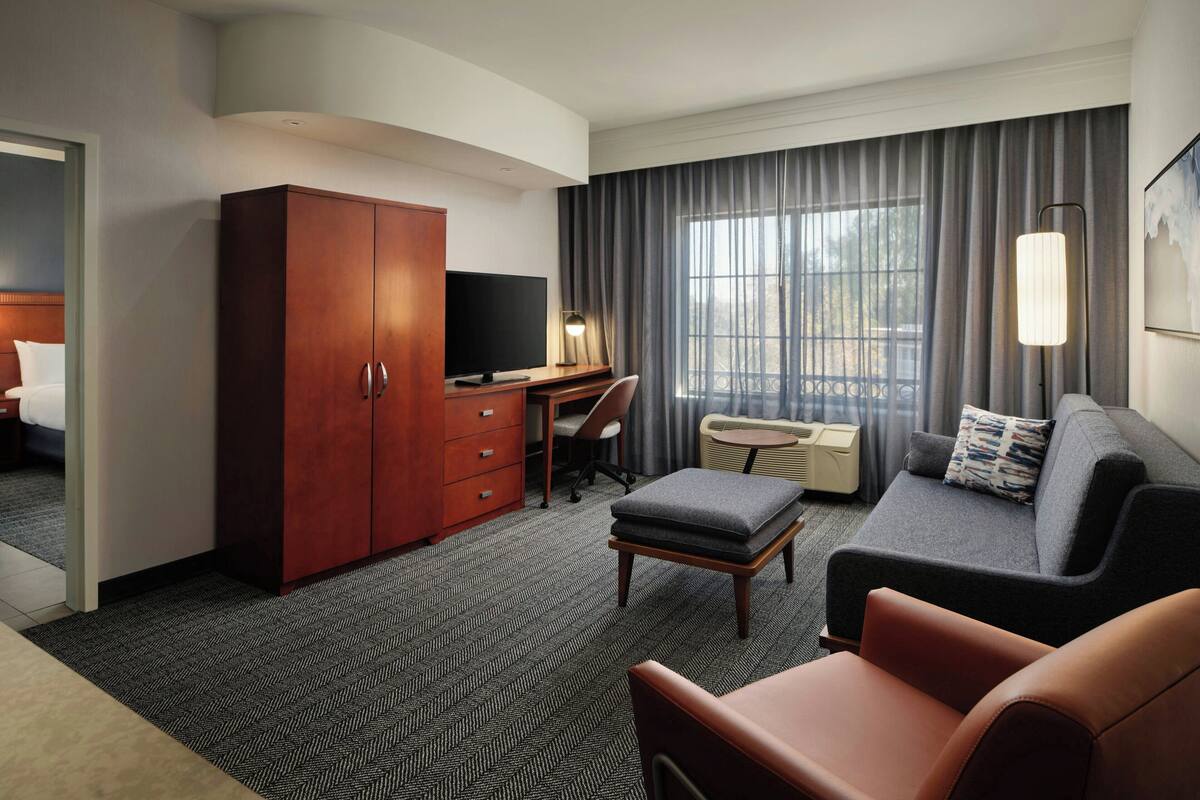 Une chambre d’hôtel avec un lit, un bureau avec une télévision, un canapé et une grande fenêtre avec des rideaux.