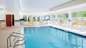 Una piscina interior con agua azul cristalina, rodeada de sillas amarillas y blancas, y grandes ventanales que ofrecen vistas del exterior.