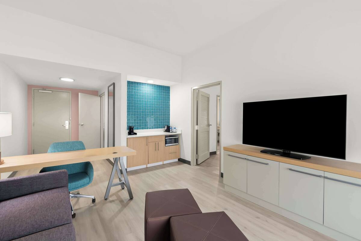 Ein modernes Hotelzimmer mit Schreibtisch aus Holz, einem blauen Sessel, einem Flachbildfernseher und einer Küchenzeile.