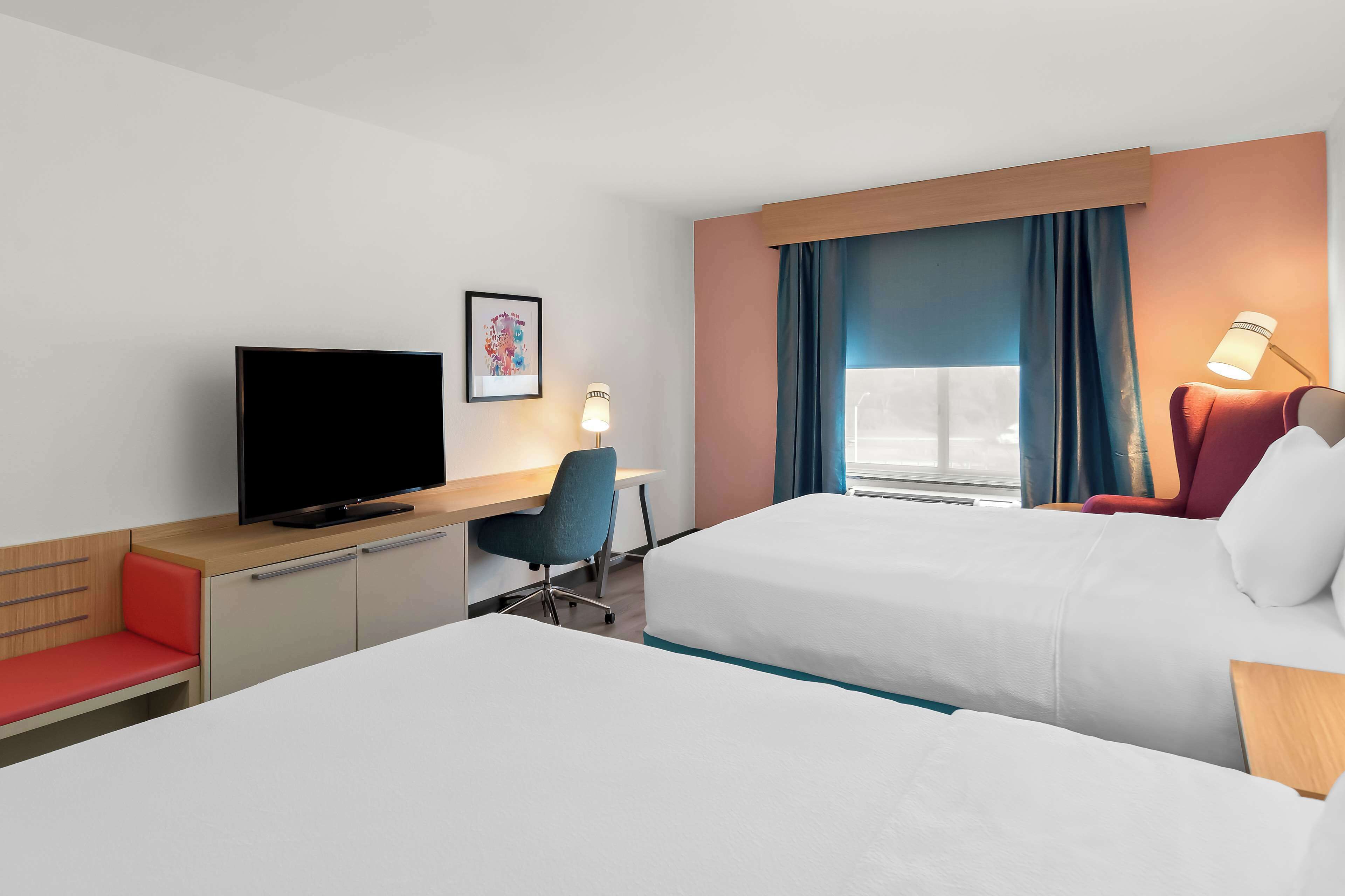 A hotel room with two beds, a desk with a TV, a chair, a lamp, and a window with curtains.