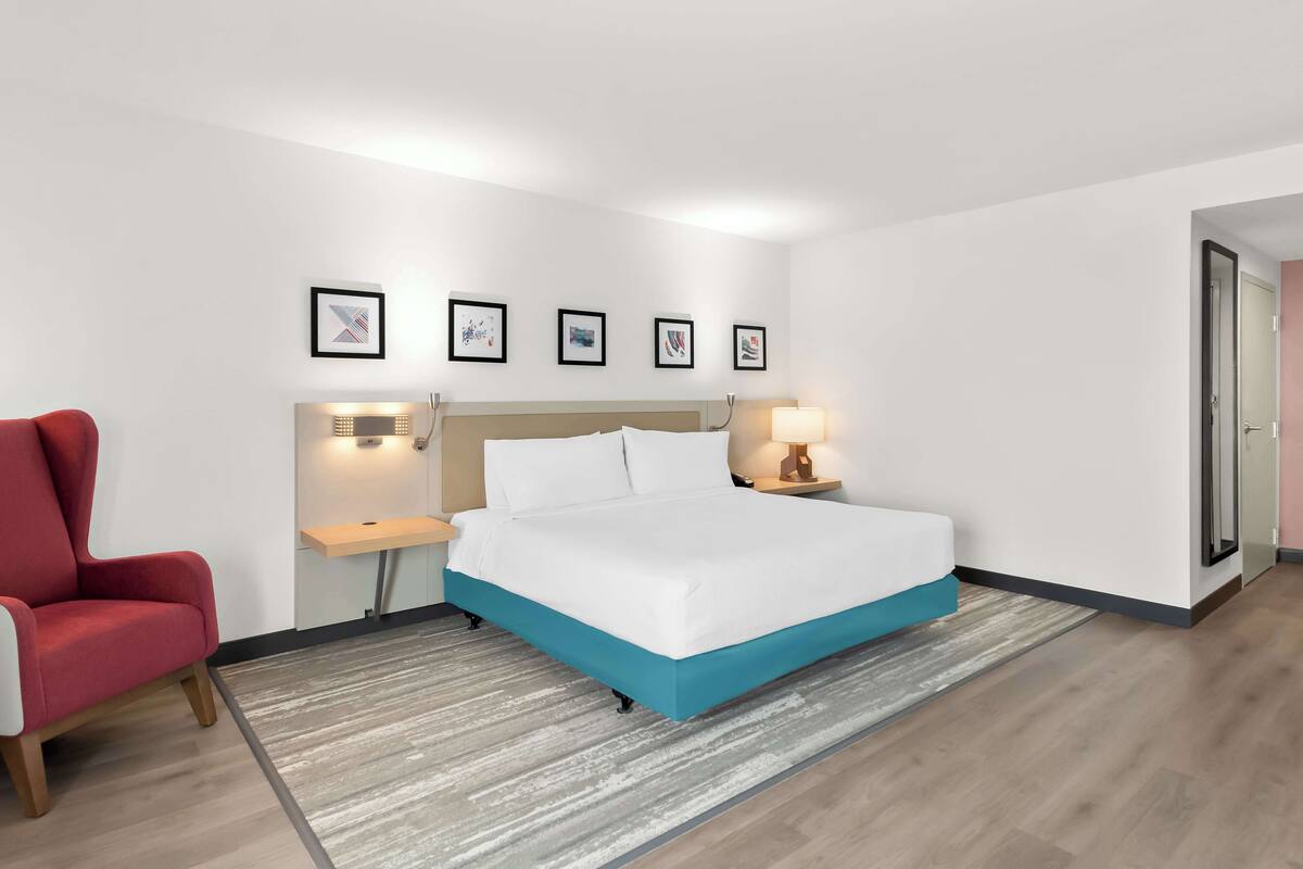 Ein modernes Hotelzimmer mit Bett, einem roten Sessel, Nachttischen mit Lampen und gerahmten Kunstwerken an der Wand.