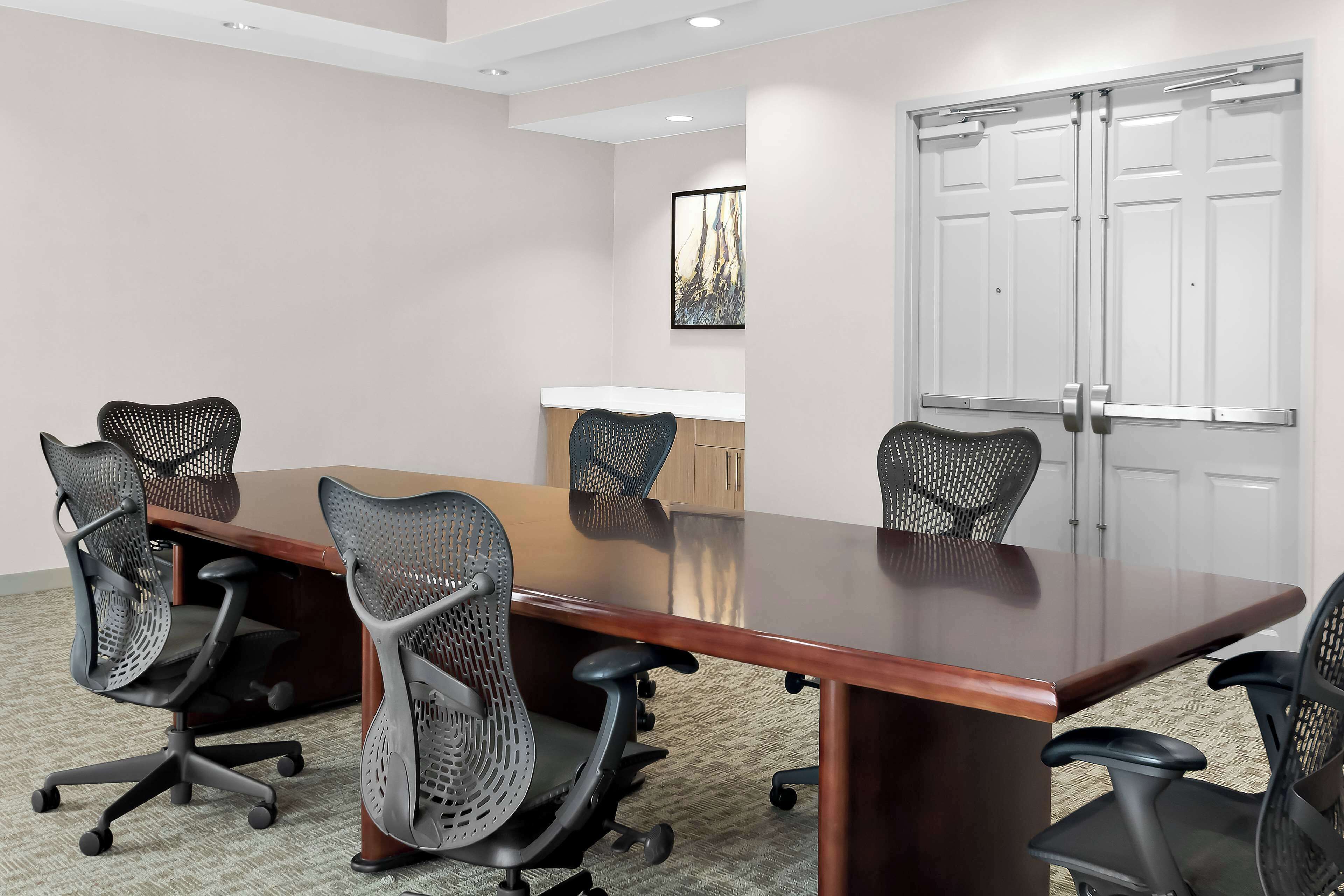 A conference room with a long table, six mesh office chairs, a whiteboard, and a door with a handle.