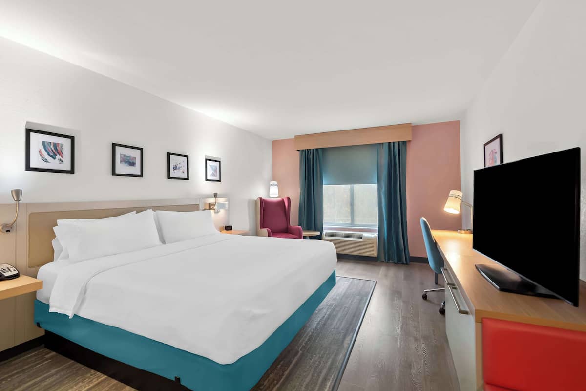 Ein Hotelzimmer mit einem großen Bett, einem Schreibtisch mit Fernseher, einem Sessel, einer Schreibtischlampe und gerahmten Bildern an der Wand.