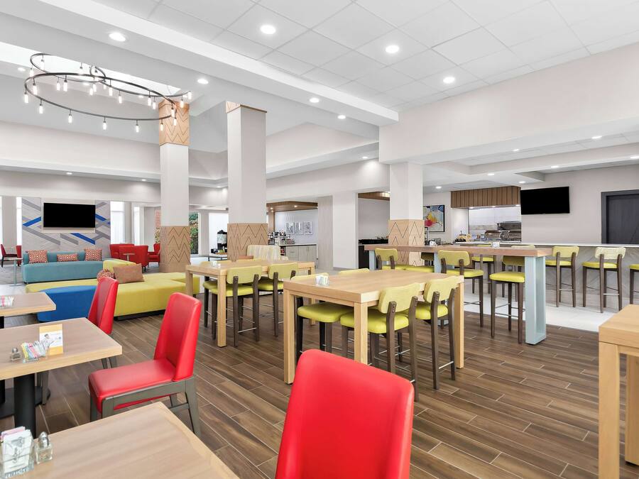 Un espace repas moderne avec plusieurs tables et chaises, une cuisine à l’arrière-plan et un téléviseur à écran plat fixé au mur.