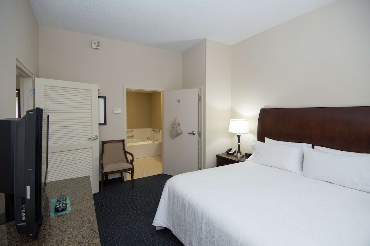 Ein Hotelzimmer mit Bett, Fernseher, Sessel, Badezimmer und Nachttisch mit Lampe.