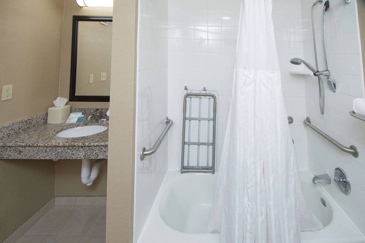 Ein Badezimmer mit Waschbecken, Spiegel, Spender für Handtücher und einer Dusche mit Haltegriff.