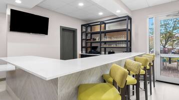 A modern bar area with a white countertop, yellow bar stools, a mounted TV, and shelves with glasses and bottles.