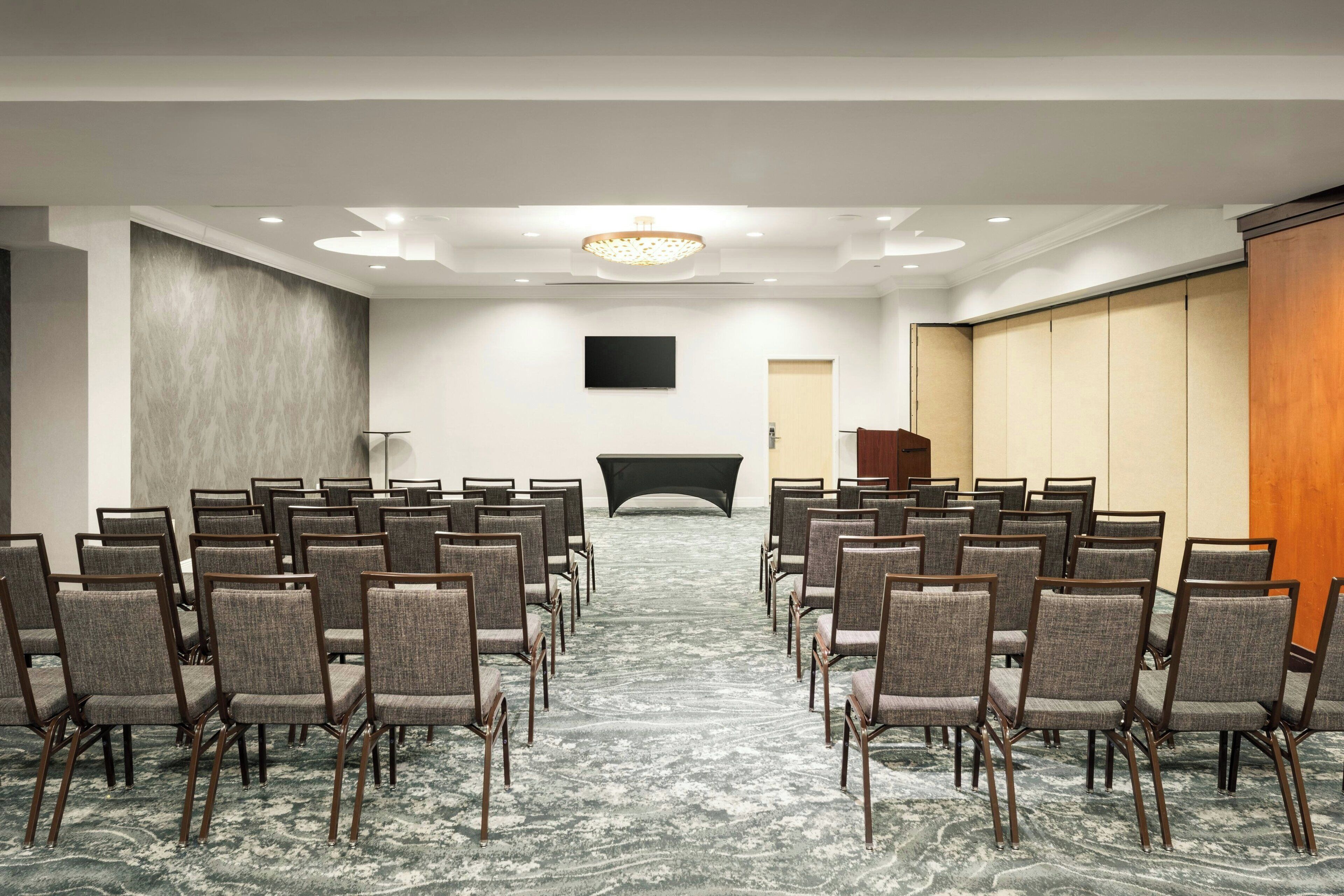 Un salón de conferencias con filas de sillas mirando hacia un escenario, un televisor de pantalla plana en la pared y un panel de madera a la derecha.