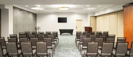 Un salón de conferencias con filas de sillas mirando hacia un escenario, un televisor de pantalla plana en la pared y un panel de madera a la derecha.