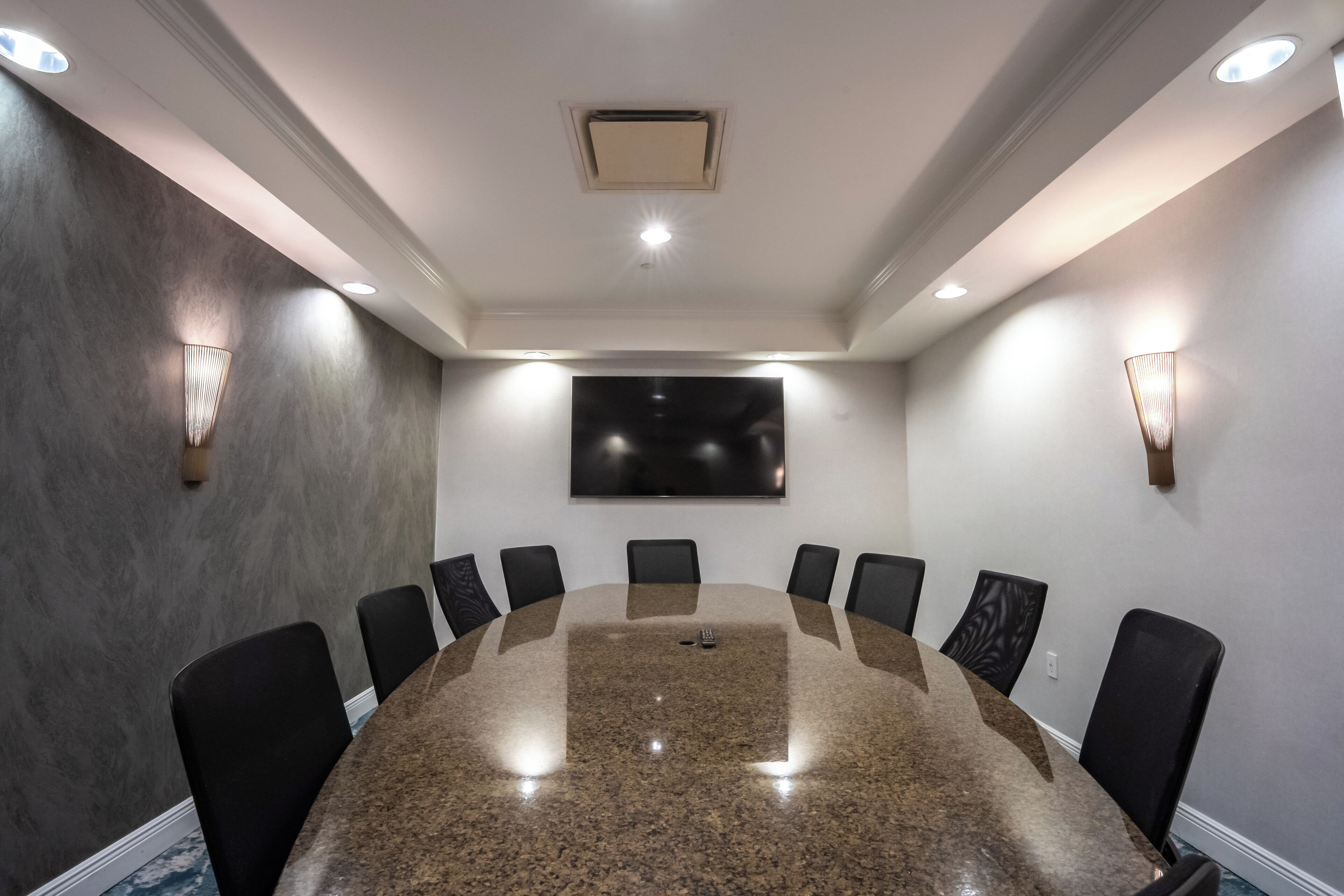 A conference room with a large oval table, black chairs, recessed ceiling lights, and a flat-screen TV on the wall.